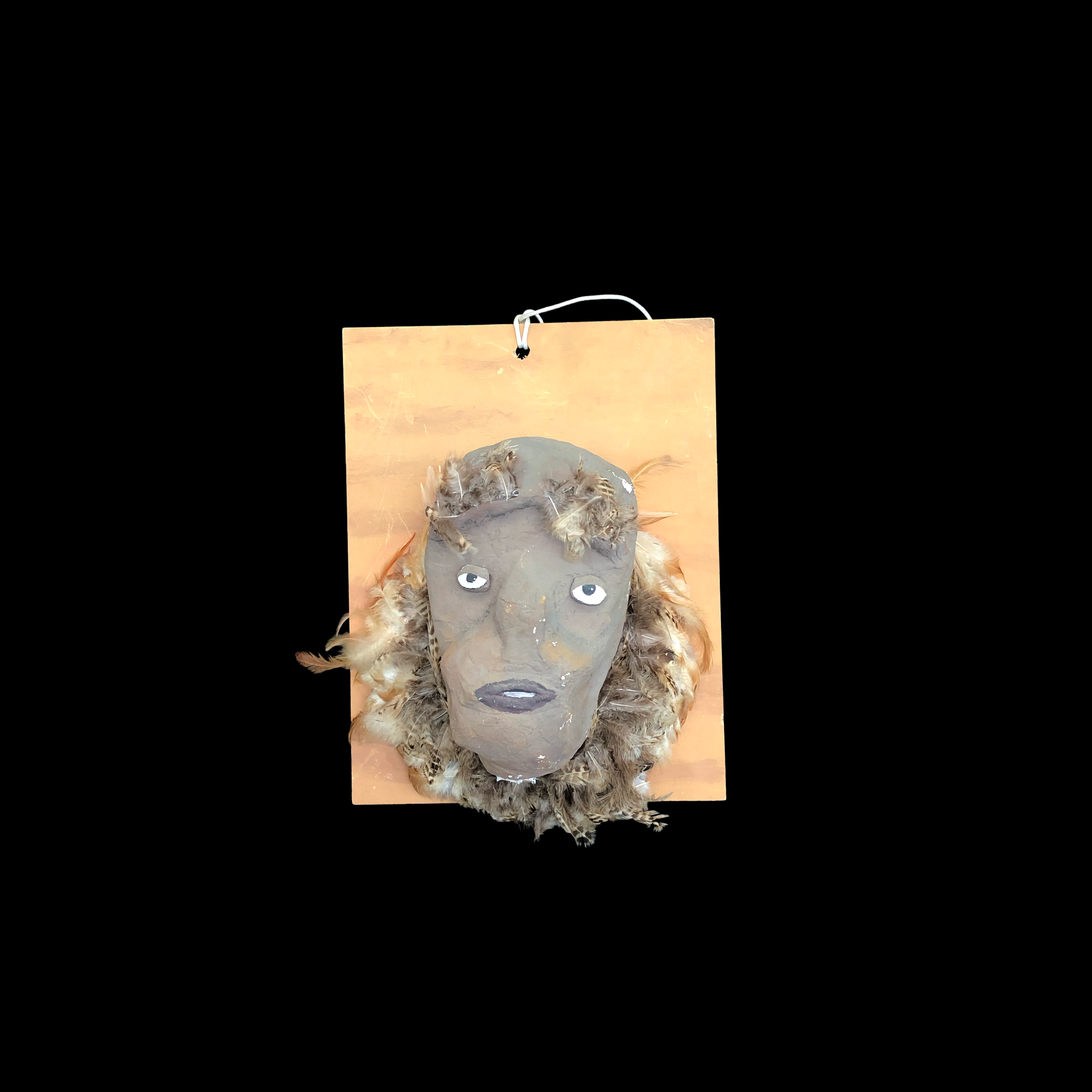 graue Maske aus Pappmaché mit Federschmuck auf Holzbrett